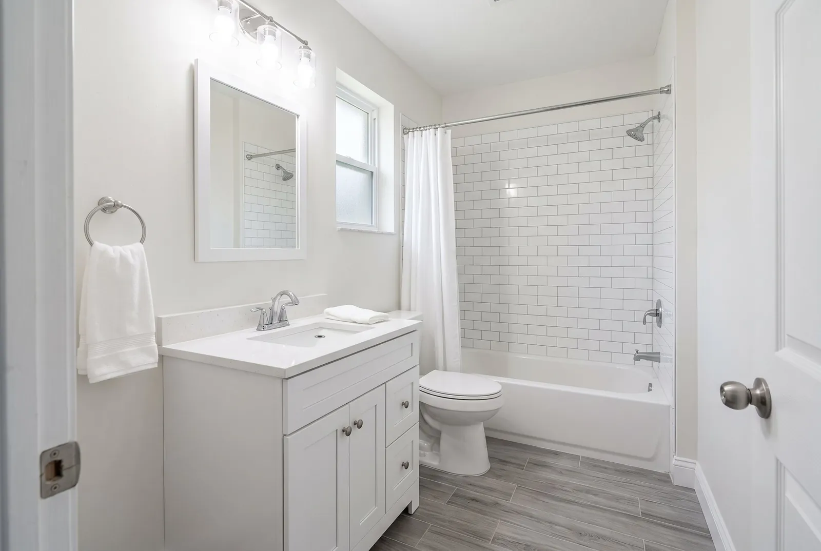 A small hall bathroom after a refresh with subway tile around the tub, a white shaker vanity and wood-look floor tile