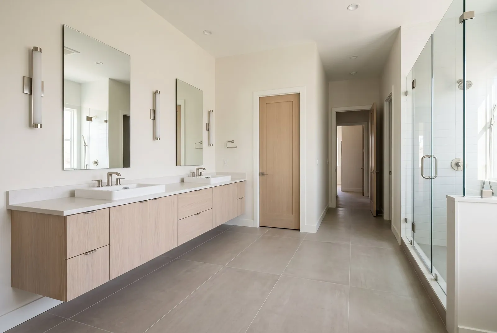 A long floating white oak double vanity with two mirrors and wall lights, and a closed wood door beyond it